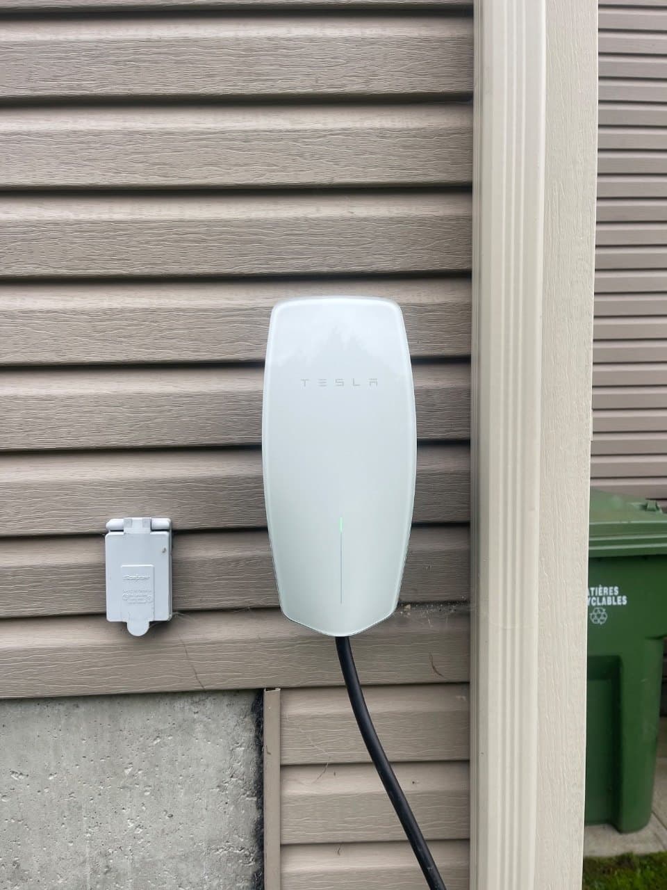 Borne de recharge Tesla installée sur le revêtement extérieur d’une maison, à côté d’une prise protégée